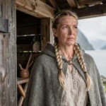 Femme viking en tenue traditionnelle nordique debout devant une maison longue en bois, regardant un fjord brumeux